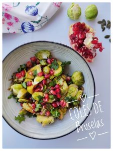 Sobre un mantel blanco se ve un bol de color gris suave. En el bol se ven unas coles de Bruselas aderecadas con granadas y cilantro. Los colores son muy vivos. Al lado del plato se ve un trapo de cocina de motivos florales, un trozo de granada, dos coles crudas y pipas de calabaza. Se trata de un plato rico en glucosinolatos para optimizar la salud físical y mental.