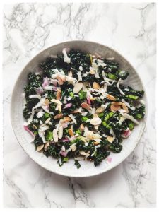 Sobre una superficie de mármol blanco se ve un plato blanco con un salteado de col kale, rábano blanco rallado, cebolla y láminas de almendra. Se trata de un plato rico en glucosinolatos para optimizar la salud física y mental