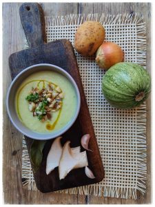 En una mesa de madera aparece una tabla de cortar con un bol. Dentro del bol hay una crema de color verde claro con frutos secos y un chorro de aceite. Al lado del bol se ven varios ingredientes un calabacín, una cebolla, una patata, setas y dos dientes de ajo