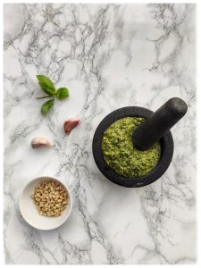 En una mesa de mármol blanco se ve un mortero de granito gris con una salsa de color verde. Al lado se ven tres ingredientes: hojas de albahaca, un pequeño bol con piñones y dos dientes de ajo