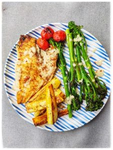 Sobre un mantel de color gris se ve un plato blanco a rayas azules. En el hay un filete de dorada a la plancha, unas patatas al horno , tres tomates cherries a la plancha y unos tallos de brocoli al vapor. Se trata de un plato rico en glucosinolatos apto para mejorar la salud física y mental