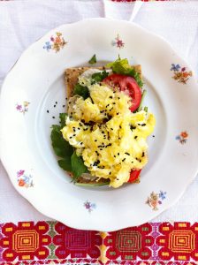 Desayuno saludable sobre mantel palestino: tostada integral con huevo revuelto, tomate fresco y hojas de rúcula (rica en glucosinolatos)  en plato blanco con diseño floral, espolvoreado con pimienta negra. Un plato óptimo para la salud física y mental.