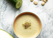 La imagen muestra un tazón blanco lleno de una sopa fría de color verde muy claro con brotes de alfalfa sobre una mesa redonda de mármol. Junto al tazón hay un pepino cortado en rodajas y al otro lado medio melón. También se ven almendras enteras y en rodajas sobre la mesa.