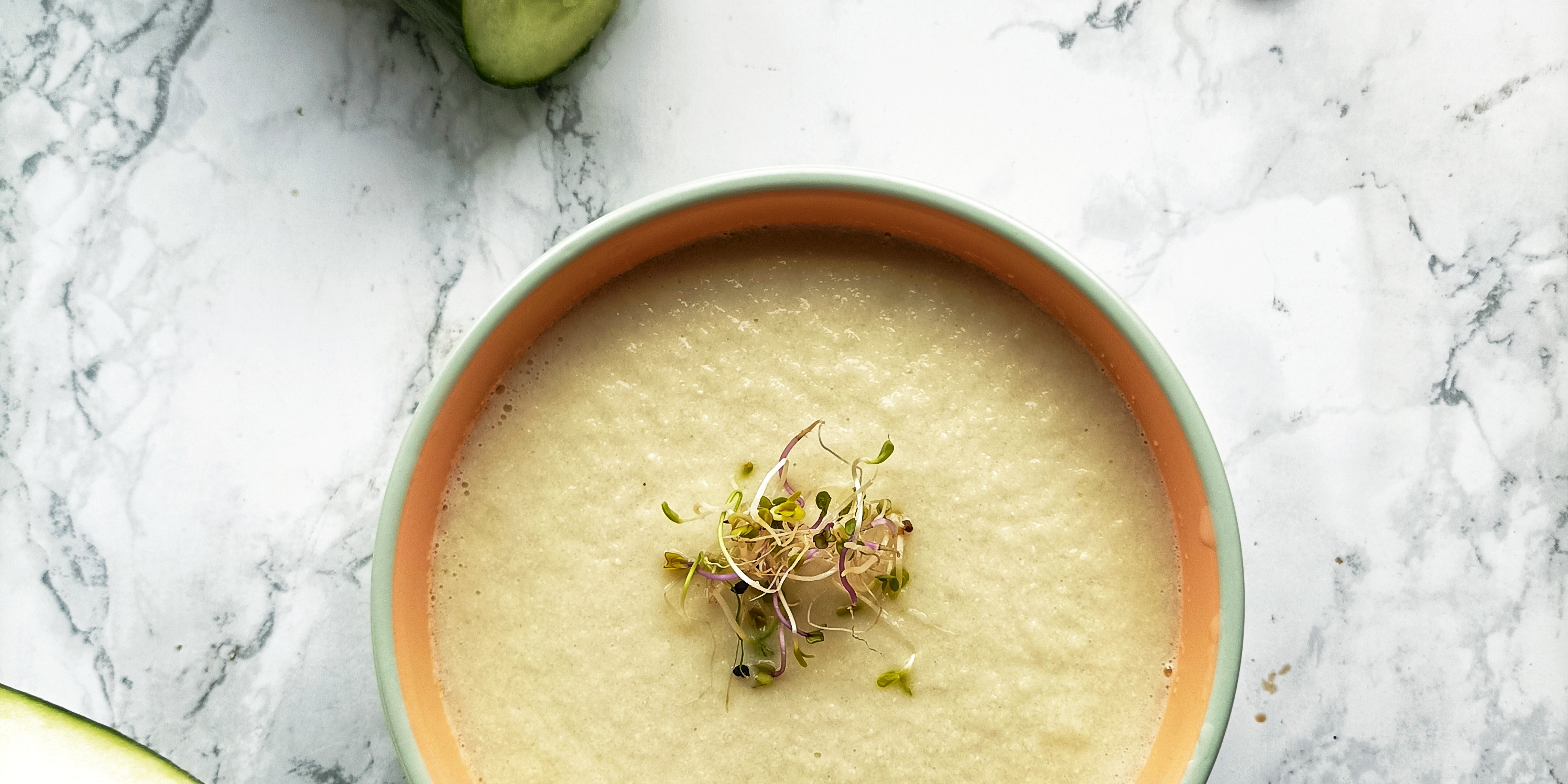 La imagen muestra un tazón blanco lleno de una sopa fría de color verde muy claro con brotes de alfalfa sobre una mesa redonda de mármol. Junto al tazón hay un pepino cortado en rodajas y al otro lado medio melón. También se ven almendras enteras y en rodajas sobre la mesa.