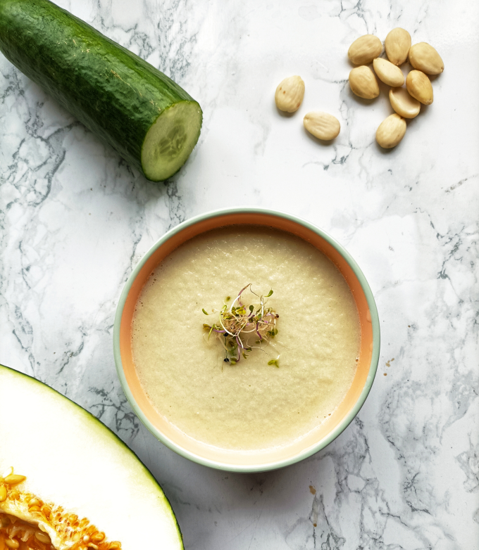 La imagen muestra un tazón blanco lleno de una sopa fría de color verde muy claro con brotes de alfalfa sobre una mesa redonda de mármol. Junto al tazón hay un pepino cortado en rodajas y al otro lado medio melón. También se ven almendras enteras y en rodajas sobre la mesa.