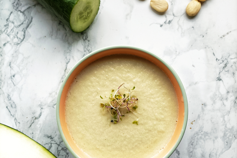 La imagen muestra un tazón blanco lleno de una sopa fría de color verde muy claro con brotes de alfalfa sobre una mesa redonda de mármol. Junto al tazón hay un pepino cortado en rodajas y al otro lado medio melón. También se ven almendras enteras y en rodajas sobre la mesa.