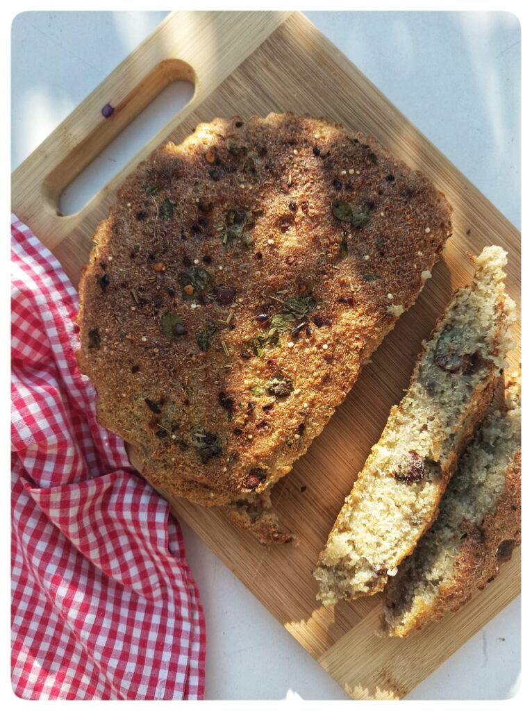 Pan de quinoa recién horneado en Air Fryer, servido en un molde de cerámica sobre una mesa de madera rústica. El pan tiene una superficie dorada con hierbas esparcidas, mostrando una textura crujiente y casera.