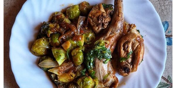 “Plato hondo con guiso mediterráneo de conejo y coles de Bruselas, acompañado de sofrito de tomate, ajo y cebolla, sobre fondo rústico de madera. Receta saludable de temporada.”