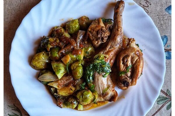 “Plato hondo con guiso mediterráneo de conejo y coles de Bruselas, acompañado de sofrito de tomate, ajo y cebolla, sobre fondo rústico de madera. Receta saludable de temporada.”