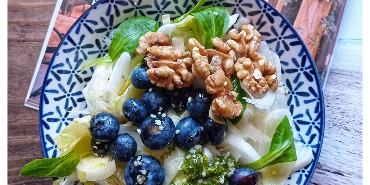 Plato de ensalada fresca con endivias, canónigos, arándanos, nueces y garbanzos sobre una mesa de madera, acompañado de una revista de viajes y luz natural. Ambiente tranquilo, sin dispositivos electrónicos a la vista.
