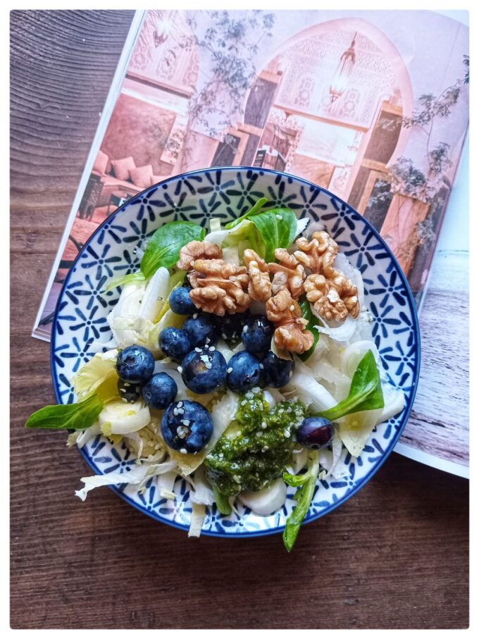 Plato de ensalada fresca con endivias, canónigos, arándanos, nueces y garbanzos sobre una mesa de madera, acompañado de una revista de viajes y luz natural. Ambiente tranquilo, sin dispositivos electrónicos a la vista.