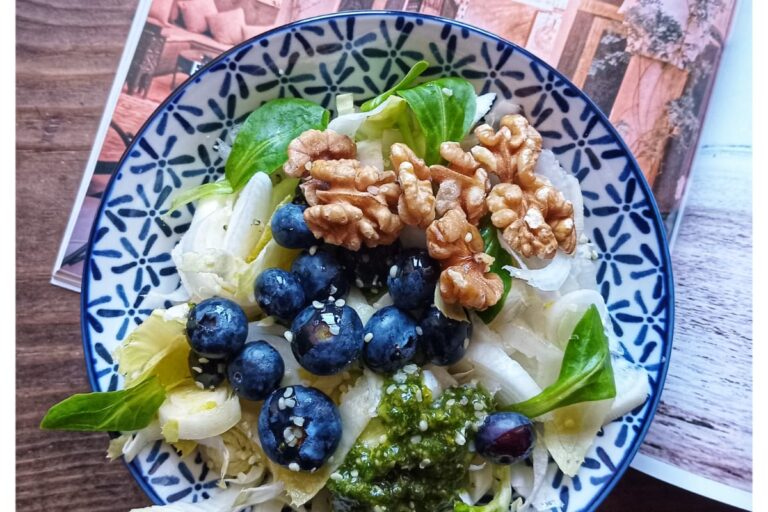 Plato de ensalada fresca con endivias, canónigos, arándanos, nueces y garbanzos sobre una mesa de madera, acompañado de una revista de viajes y luz natural. Ambiente tranquilo, sin dispositivos electrónicos a la vista.